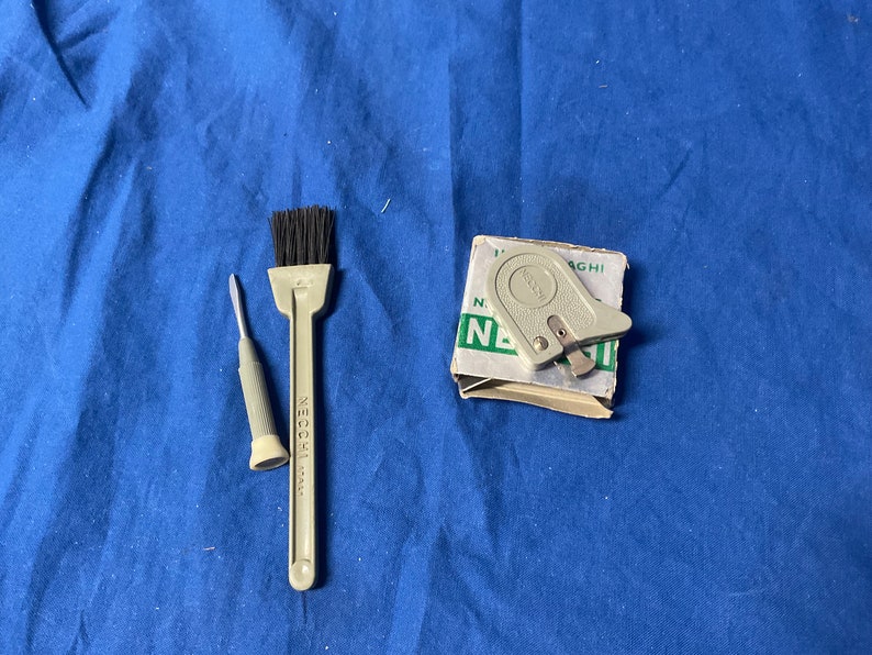 Peut inclure: Un ensemble d'outils de couture comprenant un petit tournevis gris, une brosse grise &agrave; poils noirs et un enfile-aiguille gris dans une bo&icirc;te blanche avec du texte vert qui dit "NEcchi 1 AGHI N. 1".