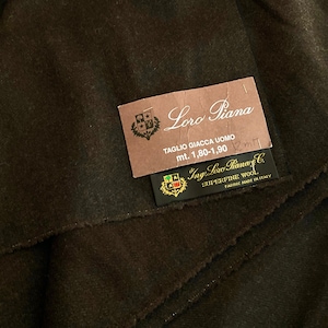 May include: Brown fabric swatch with two labels. The top label is brown with the text "Loro Piana" and "TAGLIO GIACCA UOMO mt. 1,80-1,90 m". The bottom label is black with the text "Ing. Loro Piana & C. SUPERFINE WOOL FABRIC MADE IN ITALY".