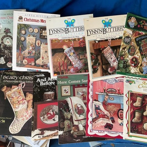 May include: A collection of 10 cross stitch patterns featuring Christmas themes. The patterns include stockings, a dollhouse, a village, and a farm. The patterns are printed on white paper with colorful illustrations.