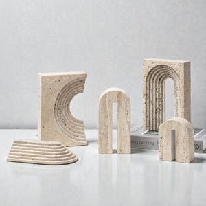 Könnte beinhalten: Eine Sammlung beigefarbener Steinskulpturen in verschiedenen geometrischen Formen. Die Stücke umfassen bogenförmige, rechteckige und halbkreisförmige Designs mit geschnitzten Details. Die Skulpturen sind auf einer weißen Oberfläche angeordnet, was eine minimalistische Ästhetik suggeriert.