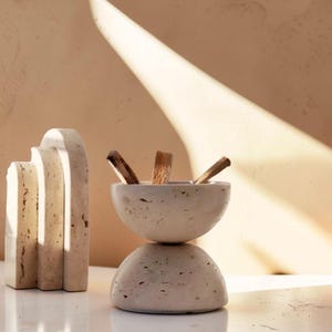 Op de afbeelding: Een lichtbeige, gestapelde stenen wierookhouder met drie wierookstokjes. De houder heeft een afgeronde basis en een komvormige bovenkant. Een bijpassende stenen sculptuur staat links.