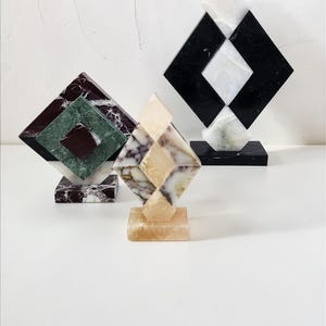May include: Three geometric marble sculptures in shades of black, white, green, and brown. Each sculpture features a diamond shape atop a square base. The sculptures are arranged on a white surface.