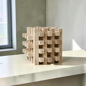 May include: A beige travertine sculpture shaped as a cube, constructed from a grid of interlocking rectangular stone bars. The natural porous texture of the light tan stone is visible across the geometric, open-frame structure of the object.