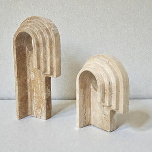 Può includere: Due sculture in pietra beige con design ad arco e stratificati. Le sculture hanno una base rettangolare e una parte superiore curva, creando una forma astratta unica. La pietra naturale presenta sottili variazioni di colore e consistenza.