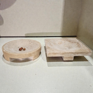 Op de afbeelding: Twee beige decoratieve stenen kommen. Eén is rond met een cirkelvormig ontwerp en de andere is vierkant met een vergelijkbaar patroon. De ronde kom heeft drie kleine voorwerpen erin. Deze kommen zijn ideaal voor het uitstallen van kleine items.
