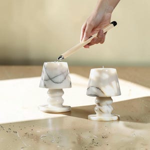 May include: Two white marble lamp-shaped candles with grey veining. A hand holds a lighter over one candle. The candles have a flared shade and a decorative base. The background is a neutral beige.