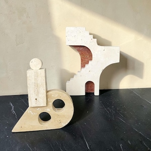 May include: Two beige travertine stone sculptures. One sculpture features a rectangular base with a circular top and two circular cutouts. The second sculpture is an architectural form with a staircase design and a reddish-brown interior arch.