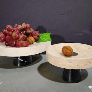 May include: Two decorative serving stands. One stand holds a bunch of red grapes, a walnut, and a green leaf. The other stand holds a single walnut. The stands have a light beige top and a black base.