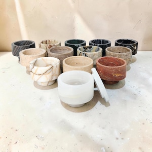 Könnte beinhalten: Eine Sammlung von Steinschalen in verschiedenen Farben und Mustern. Die Schalen sind zylindrisch mit abgerundetem Boden. Zu den Farben gehören Schwarz, Weiß, Beige, Braun und Rot. Einige Schalen haben ein marmoriertes Aussehen. Eine weiße Schale hat einen Deckel.