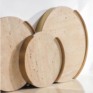 May include: A set of three circular decorative trays. The trays are made from a light beige stone material with a textured surface. Each tray has a gold-coloured metal band around the edge and a curved handle. The trays are different sizes and are stacked together.