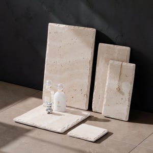 Op de afbeelding: Een verzameling van vijf rechthoekige en vierkante beige travertijn stenen platen met natuurlijke poreuze texturen en ruwe randen. Twee glazen parfumflesjes en een klein zilveren berenbeeldje staan op de grootste plaat, een gouden ketting hangt aan een andere.