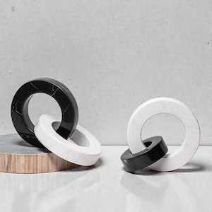 May include: Two decorative marble ring sculptures. One features black and white marble rings interlocked, the other has white and black marble rings. The sculptures are on a light grey surface with a neutral background.