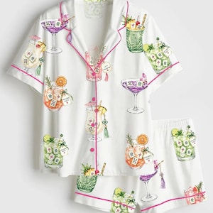 May include: A white short-sleeve button-down pajama shirt and matching shorts set featuring a colorful print of various cocktails garnished with fruit and Mahjong tiles. The garment has bright pink piping along the collar and edges.