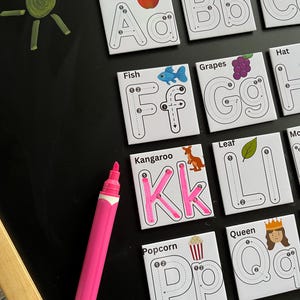 May include: A collection of white square educational tiles with letters and corresponding images, such as an apple for 'A' and a fish for 'F'. A pink marker is positioned next to the tiles on a black chalkboard. A green sun is drawn on the chalkboard.