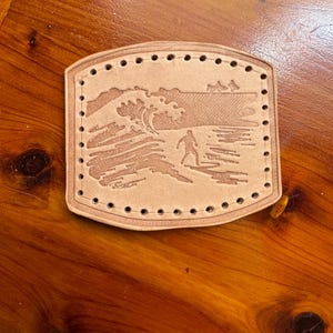 May include: A rectangular tan leather patch with rounded corners, featuring a debossed graphic of a person surfing a large wave. The patch has a series of pre-punched stitching holes along the entire perimeter, creating a border for attachment.