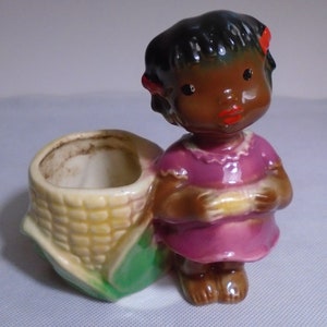 May include: A ceramic figurine of a young girl in a pink dress, holding a corn cob. The girl has dark skin, black hair with red bows, and red lips. She stands next to a cornucopia-shaped planter.