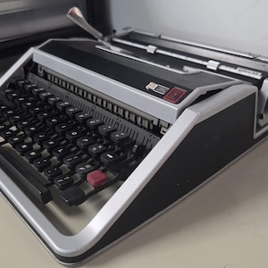 May include: A vintage typewriter with a silver and black color scheme. The keyboard has black keys with white lettering and a red return key. The typewriter has a silver carriage return lever and a black body.