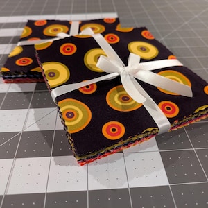 Peut inclure: Deux piles de carrés de tissu attachées avec un ruban de satin blanc. Le tissu présente un fond noir avec un motif répétitif de cercles concentriques dans des tons de jaune, orange et rouge. Les bords des carrés de tissu sont dentelés.