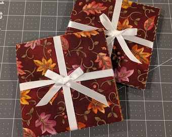 May include: Two square fabric coasters featuring a dark maroon background with a repeating pattern of autumn leaves in shades of orange, yellow, and pink, accented by brown vines and small berries, each tied with a white satin ribbon bow.