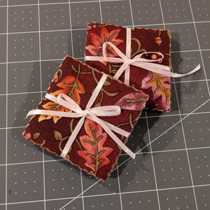 Op de afbeelding: Twee vierkante stoffen onderzetters met een donker kastanjebruine achtergrond en een herhalend patroon van oranje, roze en bruine herfstbladeren en kleine eikeltjes. Elke onderzetter is vastgebonden met een dun, lichtroze lint in een kruisvorm.