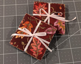 May include: Two square fabric coasters featuring a dark maroon background with a repeating pattern of orange, pink, and brown autumn leaves and small acorns. Each coaster is tied with a thin, light pink ribbon wrapped in a cross pattern.