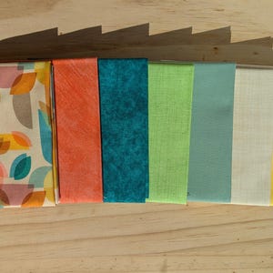 May include: A collection of folded fabric squares in various colors and patterns. The fabrics include a geometric print, solid orange, teal, lime green, seafoam, off-white, yellow, and gold. The fabrics are arranged on a wooden surface.