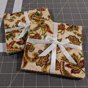 Op de afbeelding: Twee stapels vierkante stoffen onderzetters samengebonden met wit satijnen lint. De stof heeft een crèmekleurige achtergrond met een herhalend paisley-patroon in tinten rood, olijfgroen, geel en donkerbruin. De randen zijn gekarteld.