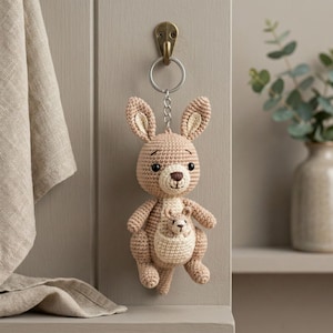 Puede incluir: Un llavero de ganchillo con forma de canguro, en tonos beige y marrón claro. El canguro tiene un pequeño en su bolsa y cuelga de un anillo de metal sujeto a un gancho de latón. El peluche hecho a mano tiene una expresión amigable.
