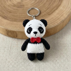 Może przedstawiać: Ręcznie robiony brelok do kluczy w kształcie pandy zrobiony na szydełku. Panda ma białe ciało, czarne ramiona, nogi i uszy oraz czerwoną muszkę. Brelok jest przymocowany do srebrnego metalowego kółka.