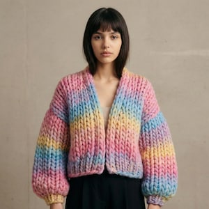 Puede incluir: Cárdigan de punto grueso en colores arcoíris, incluyendo rosa, amarillo y azul. El suéter de frente abierto tiene mangas largas y voluminosas y un ajuste relajado. La prenda está hecha de hilo grueso.