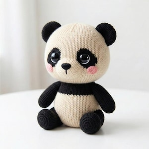 May include: A knitted panda bear plush toy. The panda has a cream-colored body with black accents on the ears, arms, and legs. It features black eyes, a black nose, and pink blush on the cheeks. The panda is sitting upright.