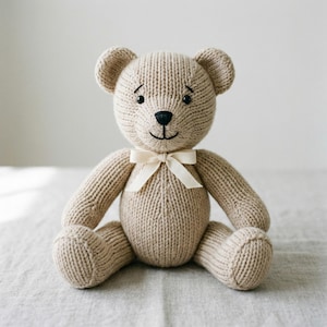 May include: A knitted teddy bear in a light tan color, with a cream-colored bow tied around its neck. The bear has a black nose and small, embroidered eyes. The bear is sitting on a neutral-colored surface.