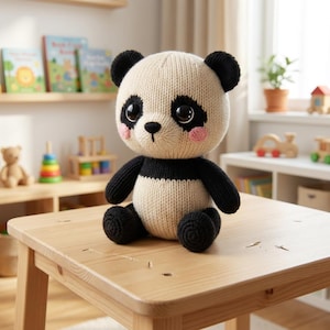 May include: A knitted panda bear toy with black and white coloring, sitting on a wooden table. The panda has black ears, eyes, and limbs, with a white face and body. Pink blush accents the cheeks. The toy is in a child's playroom.
