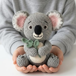 Può includere: Un koala all'uncinetto grigio fatto a mano con naso nero, orecchie rosa e bianche e occhi marroni. Il koala tiene in mano una foglia verde e ha la pancia color crema. È un peluche.