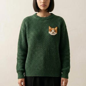 May include: A green, ribbed-knit sweater featuring a small, embroidered cat face on the chest. The cat has orange and white fur. The sweater has a crew neck and long sleeves. The bottom half of the person wearing the sweater is black.