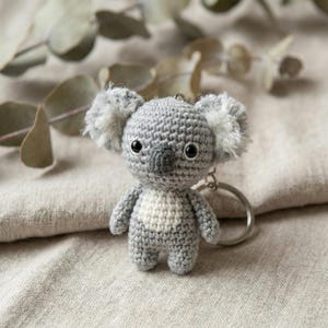 May include: A crocheted koala keychain in shades of gray and white. The koala has fluffy ears, a white chest, and black eyes. The keychain is attached to a silver metal ring. The background is a neutral linen fabric.