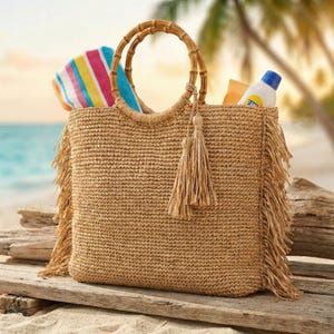 May include: A woven straw beach bag with bamboo handles and fringe detail. The bag is filled with a striped towel and sunscreen, perfect for a day at the beach. The bag is sitting on a wooden surface.