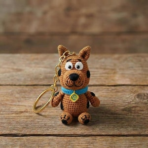 May include: Crocheted brown dog keychain with black spots, a blue collar, and a gold-colored chain and ring. The dog has large white eyes and a black nose. The keychain is sitting on a wooden surface.