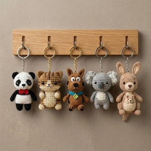 Può includere: Una collezione di cinque portachiavi a forma di animale all'uncinetto appesi a un supporto in legno. I portachiavi includono un panda, un gatto, un cane, un koala e un canguro. Ogni portachiavi ha un anello e una catena dorati.