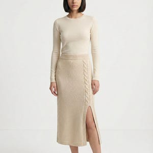Puede incluir: Top beige de manga larga y falda de punto a juego. La falda presenta un diseño de punto trenzado en el lateral y una abertura en el muslo. El conjunto está hecho de un material suave y de tonos neutros, ideal para un look casual.