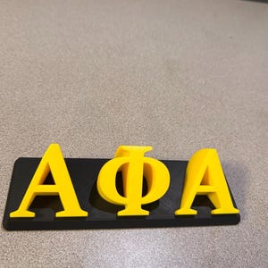 以下が含まれることがあります： 明るい黄色でギリシャ文字のアルファ、ファイ、アルファが描かれた黒い長方形のベース。文字は盛り上がっており、暗い背景の中で際立っています。このアイテムはおそらく装飾品またはサインです。