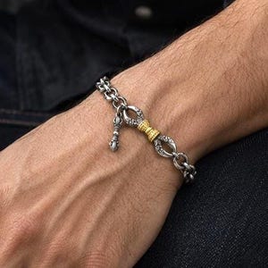 May include: A silver chain bracelet featuring oval links with an oxidized finish, accented by a central gold-toned cylindrical bead and a small dangling charm. The bracelet is secured with a clasp and rests on a wrist against dark clothing.