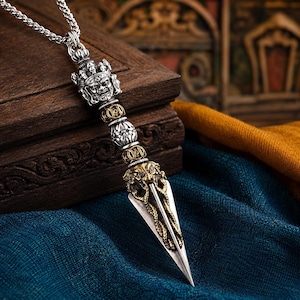 May include: A silver-toned metal pendant necklace featuring a detailed deity face at the top, followed by alternating silver and gold-toned decorative beads, and a sharp, pointed blade-like base adorned with intricate gold-toned dragon motifs.