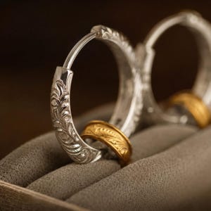 May include: A pair of silver hoop earrings featuring intricate engraved leaf patterns along the outer surface. One earring is positioned upright, while a smaller, gold-toned ring with a matching engraved leaf design rests at its base.