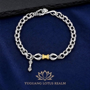 May include: A silver chain bracelet featuring engraved circular links and a central gold-toned textured barrel bead flanked by silver decorative elements. A small silver vajra charm hangs from the chain, which secures with a hook clasp.