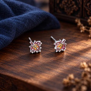 Könnte beinhalten: Ein Paar Ohrstecker in Form von Ahornblättern mit silberfarbenen Metallrändern und einer Farbverlauf-Emaillefüllung, die von tiefem Rot an den Rändern zu einem leuchtend gelben und orangen Zentrum übergeht, inklusive standardmäßiger Ohrsteckerstifte.