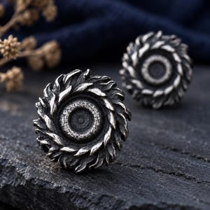 May include: A pair of circular stud earrings featuring a dark, oxidized silver finish. Each earring has a central textured disc surrounded by a ring of overlapping, flame-like or leaf-shaped metal petals, creating a layered, dimensional appearance.