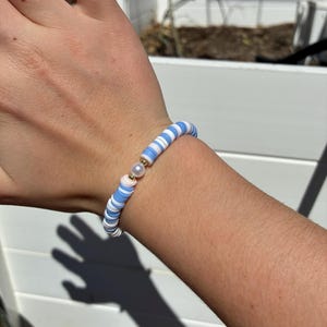 Puede incluir: Una pulsera de cuentas con cuentas en forma de disco azul claro, blanco y rosa. Una pequeña cuenta dorada y una perla adornan la pulsera. La pulsera se lleva en la muñeca.