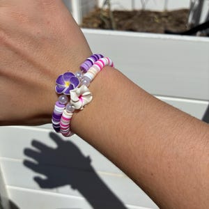 May include: A stackable bracelet set featuring a mix of colorful beads in shades of purple, pink, and white. The bracelets are adorned with flower charms and small pearl accents, creating a delicate and feminine look.