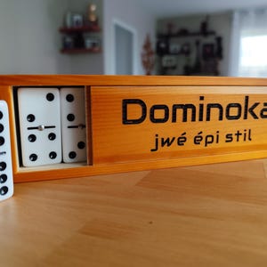Könnte beinhalten: Eine hölzerne Schiebebox mit weißen Dominosteinen mit schwarzen Punkten, auf deren Vorderseite der Text "Dominoka jwé épi stil" eingraviert ist. Ein einzelner weißer Dominostein mit schwarzen Punkten und einem Metallstift steht links daneben.